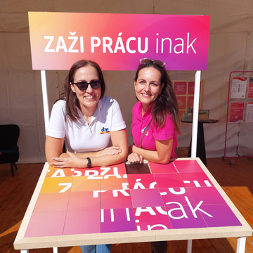 Dve usmievajúce sa ženy stoja za farebným stolom s nápisom 'ZAŽI PRÁCU inak' v jasnom, stanovom prostredí.