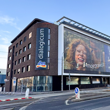Moderná tehlová budova s nápisom "dialogicum", veľkými sklenenými oknami a billboardom s usmievajúcou sa osobou. Ulica s križovatkou v popredí.