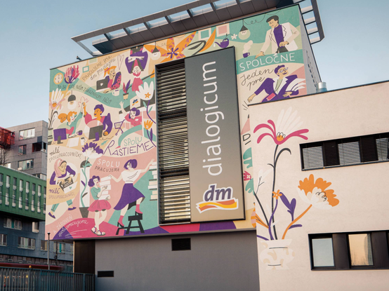 Farebná nástenná maľba na budove zobrazuje rôznych ľudí, ktorí interagujú, s textom 'dialogicum' a logom 'dm', na pozadí jasnej oblohy.