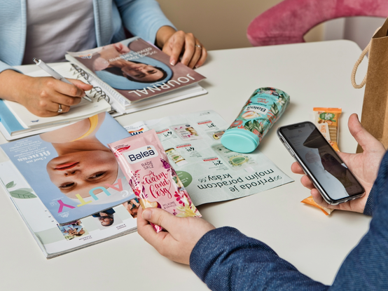 Dvaja ľudia pri stole s kozmetickými produktmi, časopismi a smartfónom. Jeden drží ružový produkt starostlivosti o pleť, zatiaľ čo druhý píše do denníka.