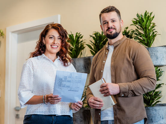 Dvaja ľudia sa usmievajú, držia dokumenty a zápisník, stoja v miestnosti s kvetináčmi a bielymi dverami v pozadí.