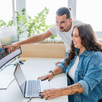 Dvaja ľudia pri stole, jeden sediaci a používajúci laptop, druhý ukazuje na počítačový monitor. Obaja sa usmievajú, v pozadí sú zelené rastliny.