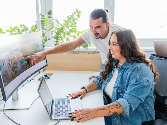 Dvaja ľudia pri stole, jeden sediaci a používajúci laptop, druhý ukazuje na počítačový monitor. Obaja sa usmievajú, v pozadí sú zelené rastliny.