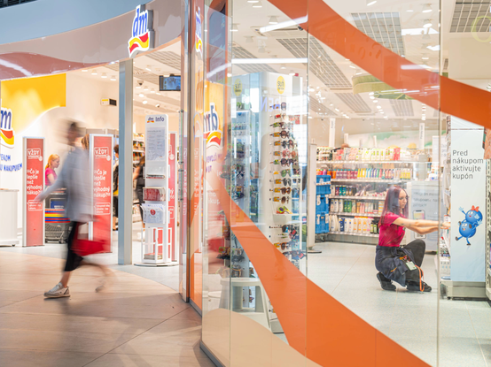 Osoba prechádza okolo jasne osvetleného obchodu so sklenenými stenami. Vo vnútri organizuje osoba police plné rôznych produktov.