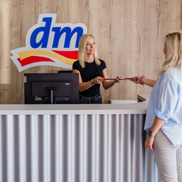 Dve ženy pri pulte na recepcii centrálneho skladu dm v Senci; jedna podáva dokument druhej. Na drevenej stene za nimi je zobrazené logo.