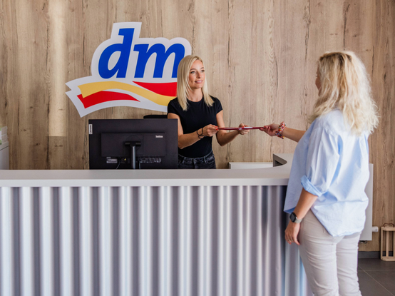 Dve ženy pri pulte na recepcii centrálneho skladu dm v Senci; jedna podáva dokument druhej. Na drevenej stene za nimi je zobrazené logo.