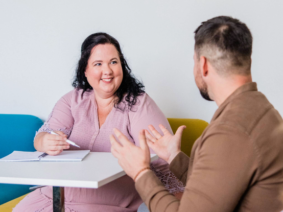 Žena v ružových šatách drží pero a usmieva sa, zatiaľ čo hovorí s mužom, ktorý gestikuluje rukami, sediacim pri stole.