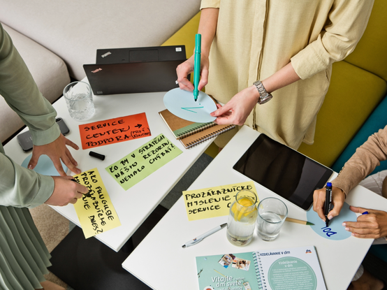 Ľudia spolupracujú pri stole s farebnými poznámkami, laptopom a nápojmi. Ruky píšu a ukazujú na rôzne papiere.
