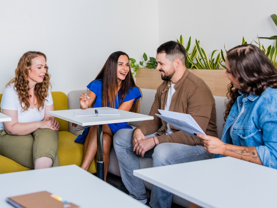 Štyria ľudia sedia a usmievajú sa počas diskusie v neformálnom kancelárskom prostredí s rastlinami v pozadí.