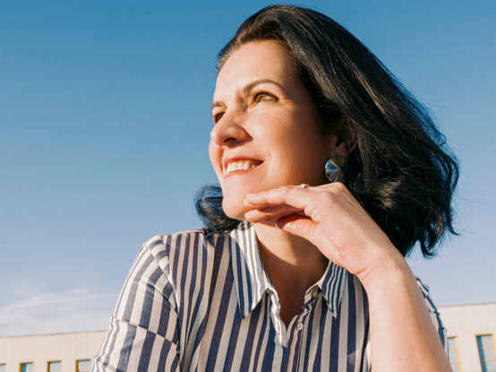 Žena s krátkymi tmavými vlasmi v pruhovanej košeli sa usmieva pod jasnou modrou oblohou. Podopiera si bradu jednou rukou.