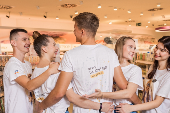 Tím piatich mladých spolupracovníkov dm v bielych tričkách sa smeje a rozpráva v obchode s kozmetikou a drogériou