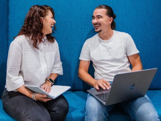 Dvaja ľudia sedia na modrom lavičke, usmievajú sa a rozprávajú. Jeden drží zápisník, druhý má laptop, obaja sú oblečení v neformálnom oblečení.