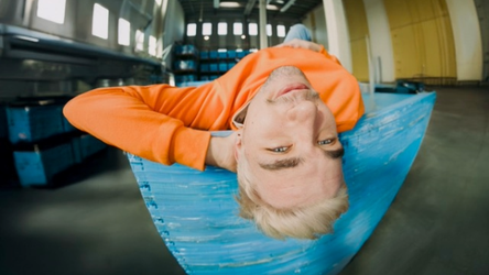 Osoba s blondfarbenými vlasmi leží hore nohami na modrom povrchu v sklade, má na sebe oranžové tričko a usmieva sa do kamery.