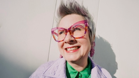 Osoba usmieva sa, nosí veľké ružové okuliare a fialovú bundu, na svetlom pozadí. Má krátke vlasy so štýlovým účesom.