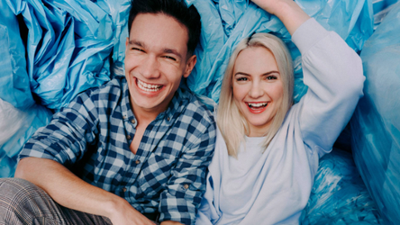 Usmievajúci sa muž a žena sedia medzi veľkými modrými taškami. Muž má na sebe károvanú košeľu a žena svetlý sveter, obaja vyzerajú šťastne.