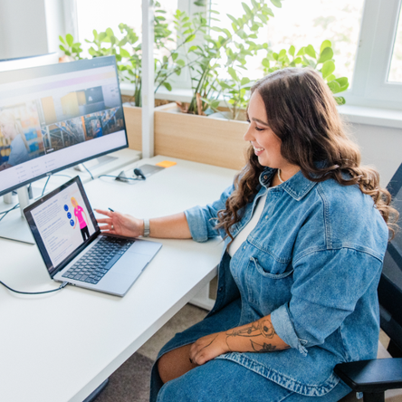 Woman in denim outfit working at a desk with a laptop and monitor, surrounded by plants, in a bright office.