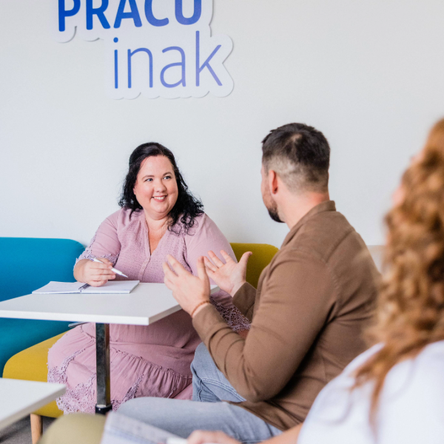 Žena a muž sedia pri stole a diskutujú v jasne osvetlenej miestnosti, za nimi je na stene nápis 'PRACUJ inak'.