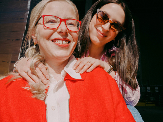 Dve ženy sa usmievajú; jedna vpredu s červenými okuliarmi a červeným kabátom, druhá vzadu v slnečných okuliaroch a ružovom oblečení, v jasnom prostredí.