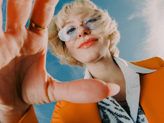 Osoba v oranžovej bunde a slnečných okuliaroch siaha smerom k fotoaparátu na pozadí modrej oblohy s hravým výrazom.